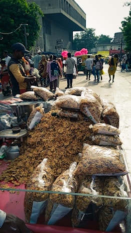 A busy street market scene with a vendor selling large bags of snack items, possibly fried crisps, displayed prominently on a table. A crowd of people is visible in the background, some walking and others gathered around different stalls. Pink balloons are seen in the distance, adding a pop of color to the bustling environment.
