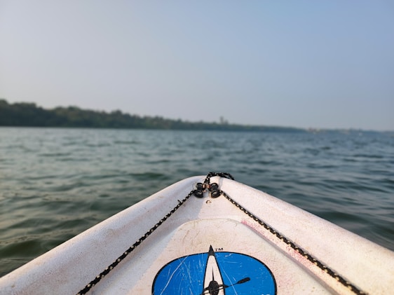 The image features the front tip of a kayak or boat with a blue logo set against a backdrop of calm water, likely a lake or river. The horizon shows a line of trees under a clear sky. The ropes on the kayak are visible, adding to the nautical theme.