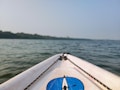 The image features the front tip of a kayak or boat with a blue logo set against a backdrop of calm water, likely a lake or river. The horizon shows a line of trees under a clear sky. The ropes on the kayak are visible, adding to the nautical theme.