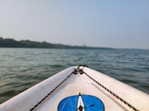 The image features the front tip of a kayak or boat with a blue logo set against a backdrop of calm water, likely a lake or river. The horizon shows a line of trees under a clear sky. The ropes on the kayak are visible, adding to the nautical theme.