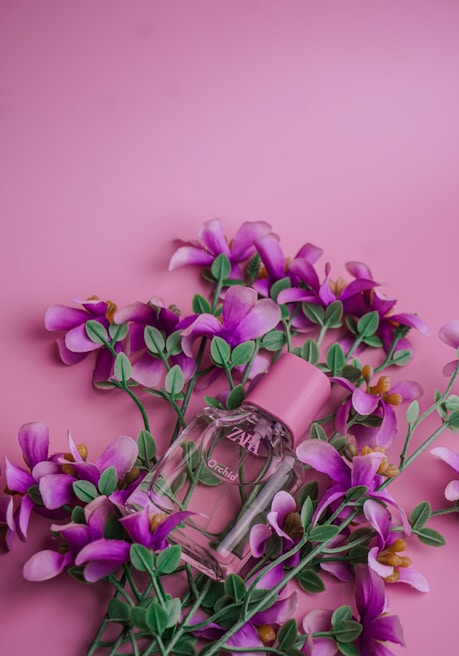A pink perfume bottle labeled 'Orchid' is surrounded by vibrant purple flowers with green leaves, set against a pink background.