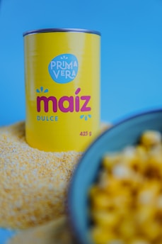 A can labeled 'Prima Vera Maíz Dulce' stands prominently against a blue background. The can is yellow with red and blue lettering, positioned on a pile of cornmeal. In the foreground, a bowl containing corn kernels is slightly out of focus.