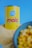 A can labeled 'Prima Vera Maíz Dulce' stands prominently against a blue background. The can is yellow with red and blue lettering, positioned on a pile of cornmeal. In the foreground, a bowl containing corn kernels is slightly out of focus.