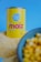 A can labeled 'Prima Vera Maíz Dulce' stands prominently against a blue background. The can is yellow with red and blue lettering, positioned on a pile of cornmeal. In the foreground, a bowl containing corn kernels is slightly out of focus.