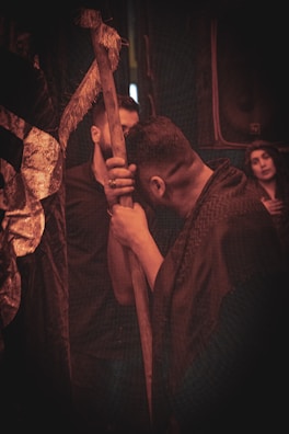 A group of people in a dimly lit setting, with one person leaning on a wooden staff. The scene has a somber and introspective tone.