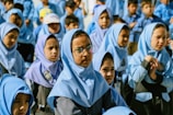 A group of young children, mostly girls, are gathered together, dressed in uniform blue hijabs and gray uniforms. One child in the foreground is wearing glasses. The children appear to be outdoors, possibly during a school event or assembly.