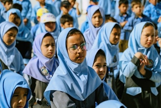 A group of young children, mostly girls, are gathered together, dressed in uniform blue hijabs and gray uniforms. One child in the foreground is wearing glasses. The children appear to be outdoors, possibly during a school event or assembly.