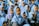 A group of young children, mostly girls, are gathered together, dressed in uniform blue hijabs and gray uniforms. One child in the foreground is wearing glasses. The children appear to be outdoors, possibly during a school event or assembly.