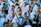 A group of young children, mostly girls, are gathered together, dressed in uniform blue hijabs and gray uniforms. One child in the foreground is wearing glasses. The children appear to be outdoors, possibly during a school event or assembly.