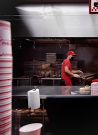 A person wearing a red uniform and cap, along with a black face mask, is working in a fast-food kitchen. There are fries in metal baskets, indicating a fryer station. A roll of paper towels is visible on the counter, with hamburger buns on a shelf below. The kitchen has a stainless steel finish and the person is handling some food items.