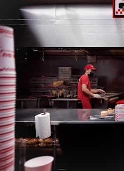 A person wearing a red uniform and cap, along with a black face mask, is working in a fast-food kitchen. There are fries in metal baskets, indicating a fryer station. A roll of paper towels is visible on the counter, with hamburger buns on a shelf below. The kitchen has a stainless steel finish and the person is handling some food items.