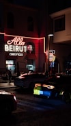 A neon-lit sign with the words 'Allo Beirut' is prominently displayed on a building. The scene takes place at night, contributing to a vibrant urban atmosphere. Several cars are visible on the cobblestone street, with one car's taillights glowing red. People can be seen inside a restaurant through the windows, adding to the bustling city vibe.