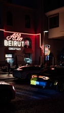 A neon-lit sign with the words 'Allo Beirut' is prominently displayed on a building. The scene takes place at night, contributing to a vibrant urban atmosphere. Several cars are visible on the cobblestone street, with one car's taillights glowing red. People can be seen inside a restaurant through the windows, adding to the bustling city vibe.