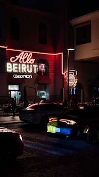 A neon-lit sign with the words 'Allo Beirut' is prominently displayed on a building. The scene takes place at night, contributing to a vibrant urban atmosphere. Several cars are visible on the cobblestone street, with one car's taillights glowing red. People can be seen inside a restaurant through the windows, adding to the bustling city vibe.