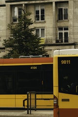 A city street scene features a stationary public bus and a tram with numbers 9811 and 4104, against a backdrop of a mid-rise residential building with balconies and windows. A tree partially obscures the building's facade, and a sign with text is visible on one of the windows.