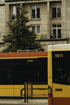 A city street scene features a stationary public bus and a tram with numbers 9811 and 4104, against a backdrop of a mid-rise residential building with balconies and windows. A tree partially obscures the building's facade, and a sign with text is visible on one of the windows.