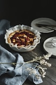 A rustic baked fruit tart is placed on a dark wooden table. The tart has a golden crust and contains slices of apples or pears topped with dark berries. It sits in a white fluted dish lined with baking paper. To the right, there are a couple of stacked white plates with forks on top, and a speckled, small plate on the edge of the image. A gray-blue linen cloth is elegantly draped to the left with delicate white dried flowers scattered across it.