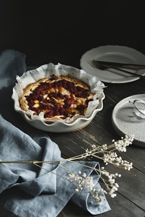 A rustic baked fruit tart is placed on a dark wooden table. The tart has a golden crust and contains slices of apples or pears topped with dark berries. It sits in a white fluted dish lined with baking paper. To the right, there are a couple of stacked white plates with forks on top, and a speckled, small plate on the edge of the image. A gray-blue linen cloth is elegantly draped to the left with delicate white dried flowers scattered across it.