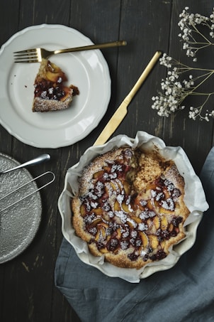 A rustic fruit tart dusted with powdered sugar sits in a ceramic dish lined with parchment paper. A piece has been sliced and placed on a white plate with a golden fork. The background features a wooden table and a blue-gray cloth napkin, while sprigs of white flowers add a decorative touch.