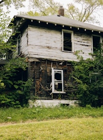 An old, abandoned house with broken windows surrounded by overgrown weeds.