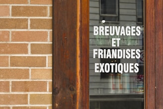A wooden-framed window displaying the text 'BREUVAGES ET FRIANDISES EXOTIQUES' in white, with a stone brick wall beside it. The reflection of a building is visible on the glass.
