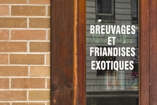 A wooden-framed window displaying the text 'BREUVAGES ET FRIANDISES EXOTIQUES' in white, with a stone brick wall beside it. The reflection of a building is visible on the glass.