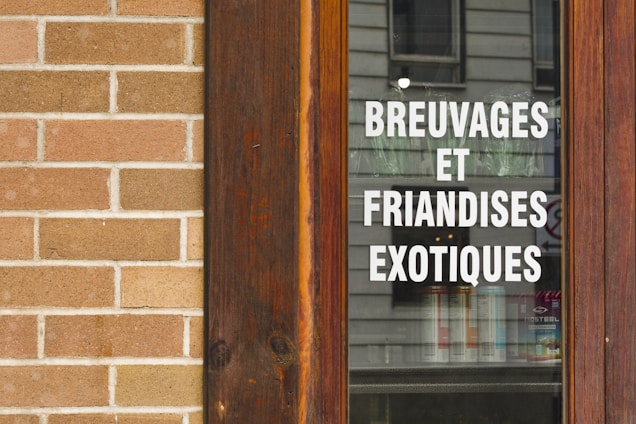 A wooden-framed window displaying the text 'BREUVAGES ET FRIANDISES EXOTIQUES' in white, with a stone brick wall beside it. The reflection of a building is visible on the glass.
