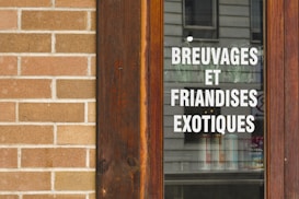 A wooden-framed window displaying the text 'BREUVAGES ET FRIANDISES EXOTIQUES' in white, with a stone brick wall beside it. The reflection of a building is visible on the glass.
