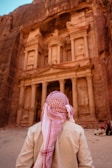 A person wearing a traditional Middle Eastern keffiyeh stands in front of a grand ancient stone structure carved into a rock face. The structure features ornate columns and intricate architectural details, suggesting historical significance.