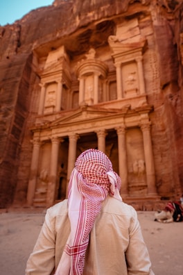 A person wearing a traditional Middle Eastern keffiyeh stands in front of a grand ancient stone structure carved into a rock face. The structure features ornate columns and intricate architectural details, suggesting historical significance.