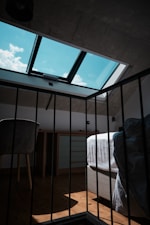 A dimly lit loft room with a skylight window allowing sunlight to stream in, casting shadows on the wooden floor. The room contains a bed with white sheets, a chair, and a dresser. The ceiling and some walls are made of exposed concrete, giving it an industrial feel.