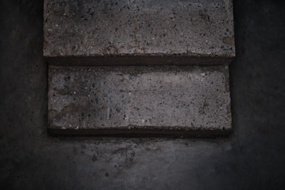 Two concrete steps with a rough texture appear worn and slightly uneven. The surface contains small pebbles and aggregate, giving it a speckled appearance. The surrounding area is a similar concrete material, darker due to shadows and lighting.