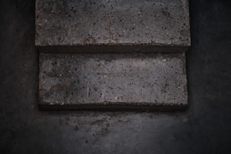 Two concrete steps with a rough texture appear worn and slightly uneven. The surface contains small pebbles and aggregate, giving it a speckled appearance. The surrounding area is a similar concrete material, darker due to shadows and lighting.