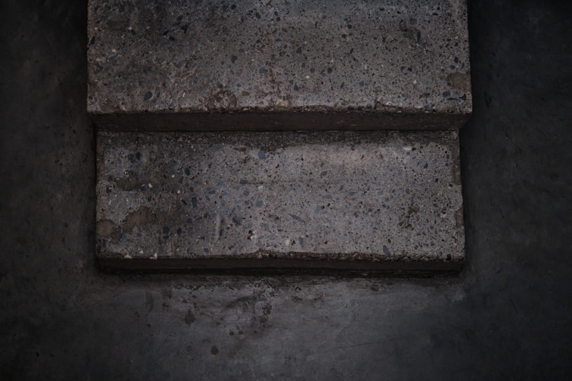 Two concrete steps with a rough texture appear worn and slightly uneven. The surface contains small pebbles and aggregate, giving it a speckled appearance. The surrounding area is a similar concrete material, darker due to shadows and lighting.