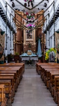 A beautifully decorated church interior featuring wooden pews aligned neatly on either side of a central aisle. The altar is adorned with flowers, greenery, and religious statues, including a central figure dressed in robes. Festive garlands and colorful ornaments hang from above, enhancing the sacred atmosphere.