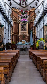 A beautifully decorated church interior featuring wooden pews aligned neatly on either side of a central aisle. The altar is adorned with flowers, greenery, and religious statues, including a central figure dressed in robes. Festive garlands and colorful ornaments hang from above, enhancing the sacred atmosphere.