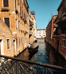 A serene canal flanked by rustic buildings with colorful facades, featuring a gondola maneuvered by a gondolier. The sunlight casts warm tones on the architecture, highlighting the intricate details of Venetian design. Shadows play across the water, adding depth and contrast to the calm scene.
