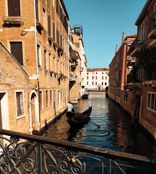 A serene canal flanked by rustic buildings with colorful facades, featuring a gondola maneuvered by a gondolier. The sunlight casts warm tones on the architecture, highlighting the intricate details of Venetian design. Shadows play across the water, adding depth and contrast to the calm scene.