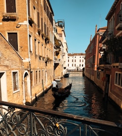 A serene canal flanked by rustic buildings with colorful facades, featuring a gondola maneuvered by a gondolier. The sunlight casts warm tones on the architecture, highlighting the intricate details of Venetian design. Shadows play across the water, adding depth and contrast to the calm scene.