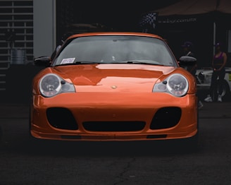 An orange sports car is parked in front of a garage or workshop. The car has sleek lines and large headlights, giving it a modern and high-performance appearance. In the background, there are people standing next to another vehicle, with some signage visible on a canopy.