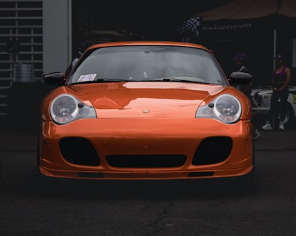 An orange sports car is parked in front of a garage or workshop. The car has sleek lines and large headlights, giving it a modern and high-performance appearance. In the background, there are people standing next to another vehicle, with some signage visible on a canopy.