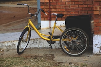 A yellow tricycle is parked on a paved area near a brick wall. The tricycle has a back carrying compartment or basket, supported by two rear wheels. The background includes a tiled platform and some dirt ground beside it.
