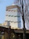 A modern high-rise building under construction with a distinctive stacked design. The structure features multiple tiers of glass facades and protruding sections. A crane is visible at the top of the building, indicating ongoing construction. The base of the building is adjacent to a brick structure with lit archways and a sign that reads 'MATRIX'. Bare trees frame the view in the foreground.
