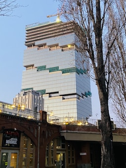 A modern high-rise building under construction with a distinctive stacked design. The structure features multiple tiers of glass facades and protruding sections. A crane is visible at the top of the building, indicating ongoing construction. The base of the building is adjacent to a brick structure with lit archways and a sign that reads 'MATRIX'. Bare trees frame the view in the foreground.