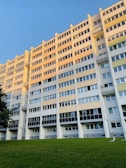 A large multi-story apartment building with multiple rows of windows reflecting the evening sunlight. The structure is painted in shades of beige, gray, and white. In front of the building, there is a well-maintained grassy lawn with two electric scooters lying near the edge. A clear, blue sky is visible above the building.