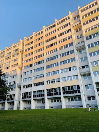 A large multi-story apartment building with multiple rows of windows reflecting the evening sunlight. The structure is painted in shades of beige, gray, and white. In front of the building, there is a well-maintained grassy lawn with two electric scooters lying near the edge. A clear, blue sky is visible above the building.