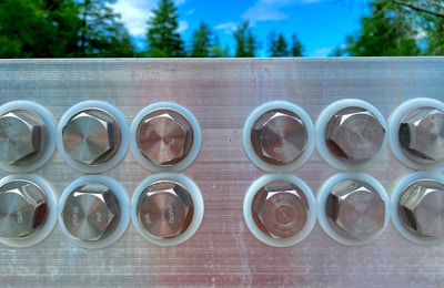A close-up of eight metallic hexagonal bolts arranged in two rows against a reflective surface. In the background, there are lush green trees and a vivid blue sky.
