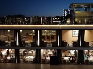 The image captures a multi-story cruise ship or building at night with a modern and elegant interior. The upper floor shows a bar with stools and tables, while the lower floor is a dining area with people seated at neatly set tables. The scene is well-lit, and the sky is dark, indicating evening or nighttime.