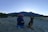 A large backpack with camping gear sits on a rocky landscape at dusk, with a German Shepherd dog sitting beside it. In the background, a mountain range under a clear sky creates a serene, natural setting.