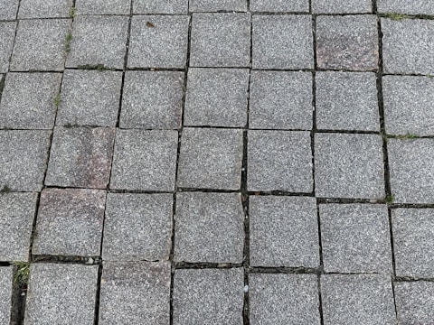 A pattern of square gray stone tiles laid out on the ground, with some gaps filled with small patches of grass. The surface texture of the tiles appears rough and slightly weathered.